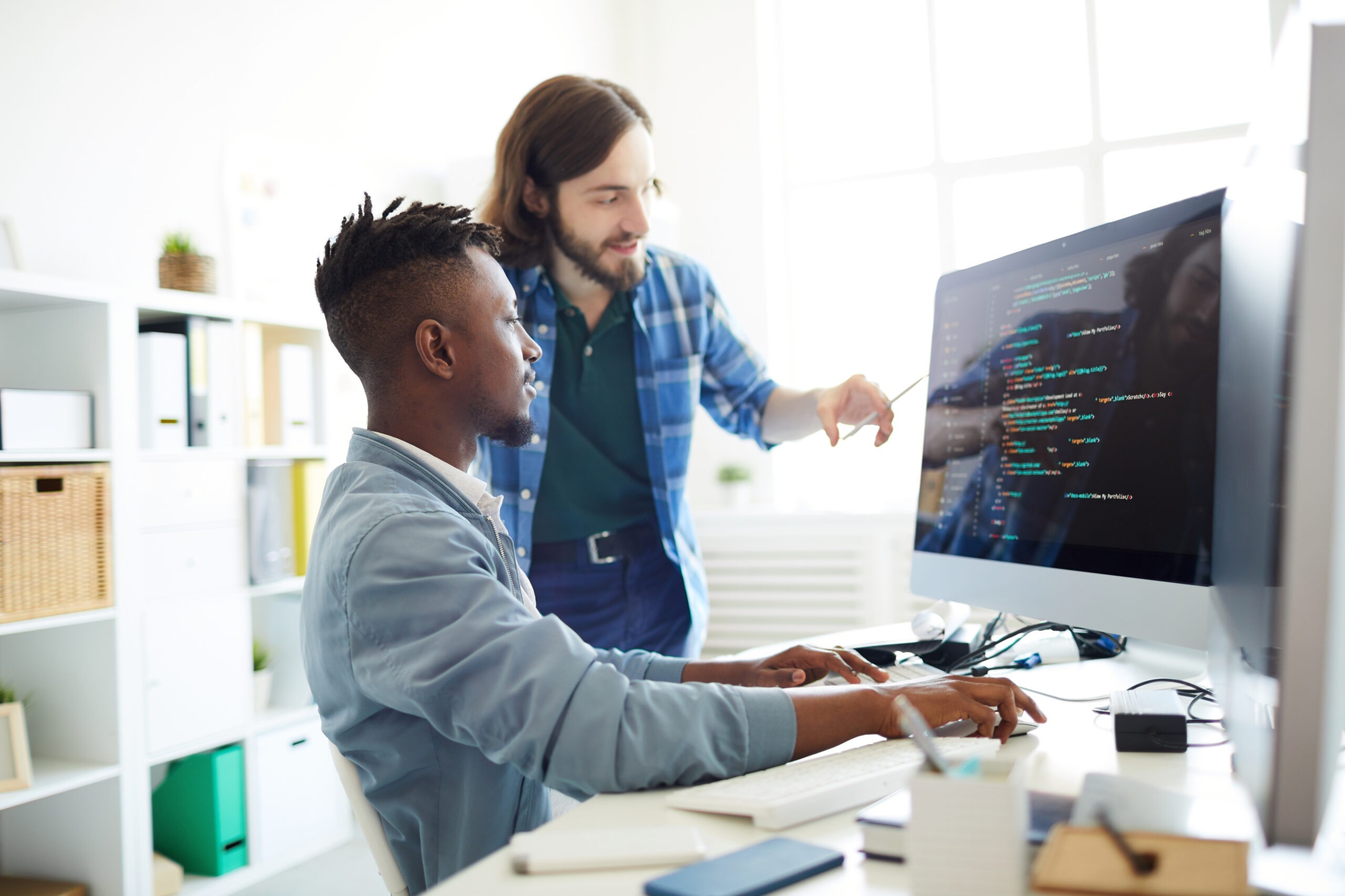 Positive skilled young multiethnic coders in casual clothing discussing computer language: smiling bearded man pointing at computer monitor while explaining web code to African colleague in office; Shutterstock ID 1145542748; Purchase Order: comps; Job: ; Client/Licensee: ; Other: